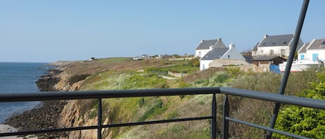 Un balcón con una silla de mimbre, con vista a un pueblo costero con casas y acantilados.