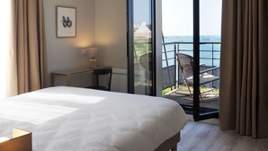 A hotel room with a large bed, a desk, a chair, a window with a view of the sea, and a balcony with outdoor seating.
