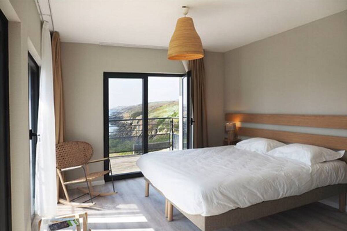 A bedroom with a large bed, a chair, a small table, and a view of the outdoors through a sliding door.