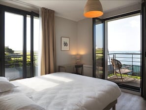 A bedroom with a large bed, a desk, and a chair, with a view of the sea through a glass door.
