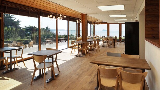 A spacious restaurant with wooden tables and chairs, large windows offering a view of the sea, and a modern ceiling design.