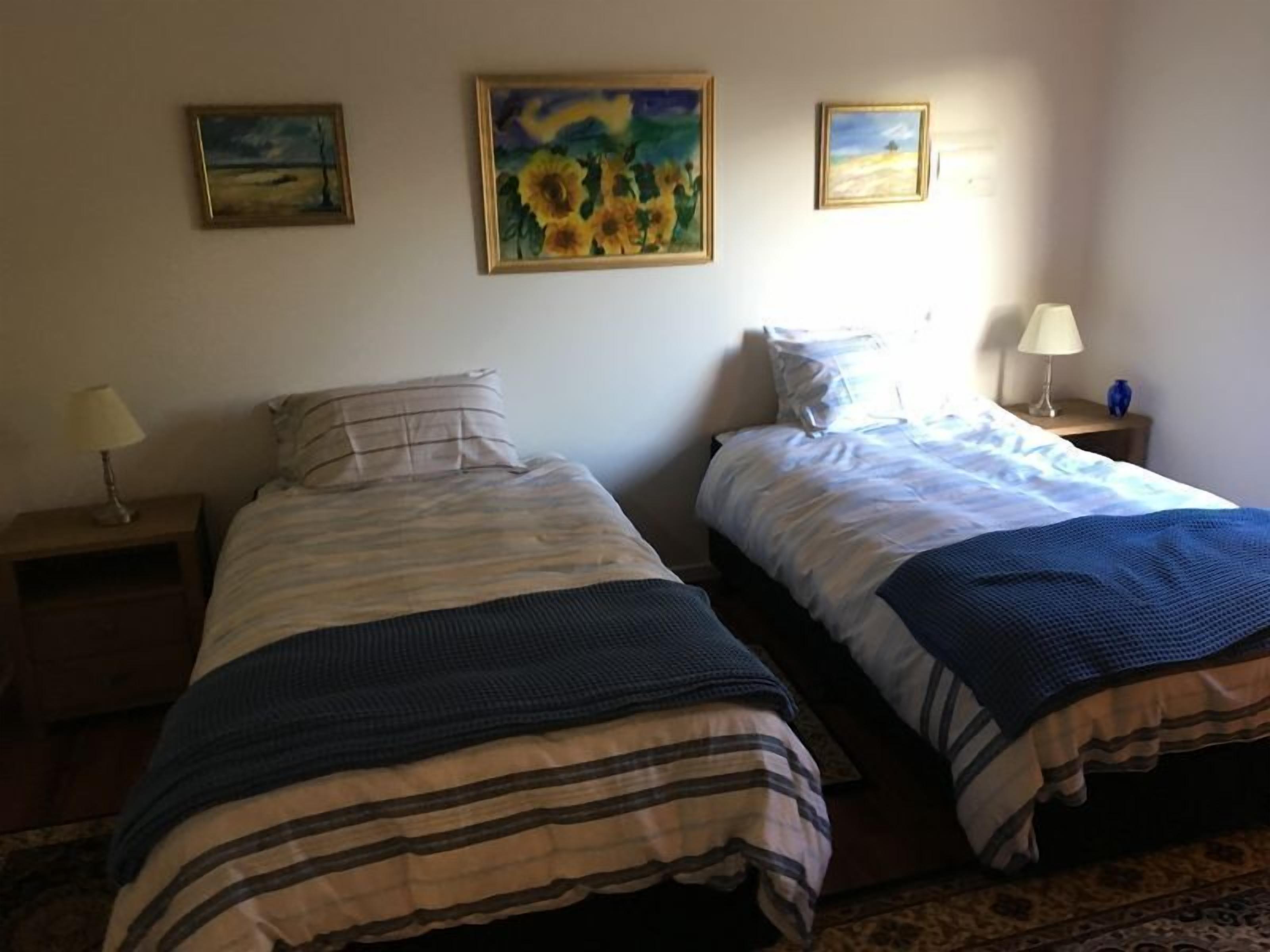 Una habitación con dos camas, cada una con una colcha a rayas, una mesita de noche con una lámpara y un cuadro en la pared.