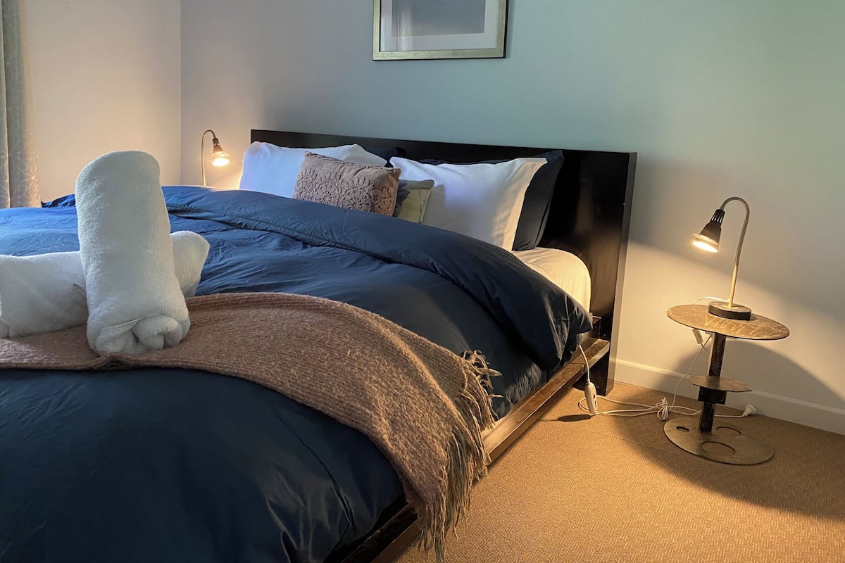 A neatly made bed with a blue and brown duvet, a white towel, and a framed picture on the wall.