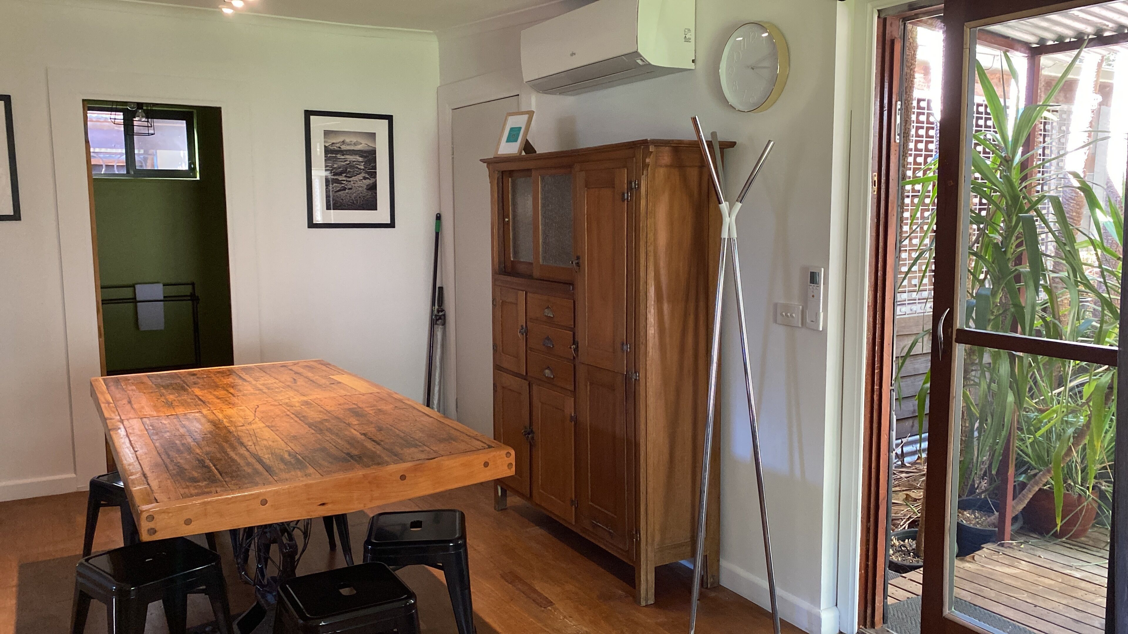 A room with a wooden table, two black stools, a wooden cabinet, and a door leading to an outdoor area with plants.