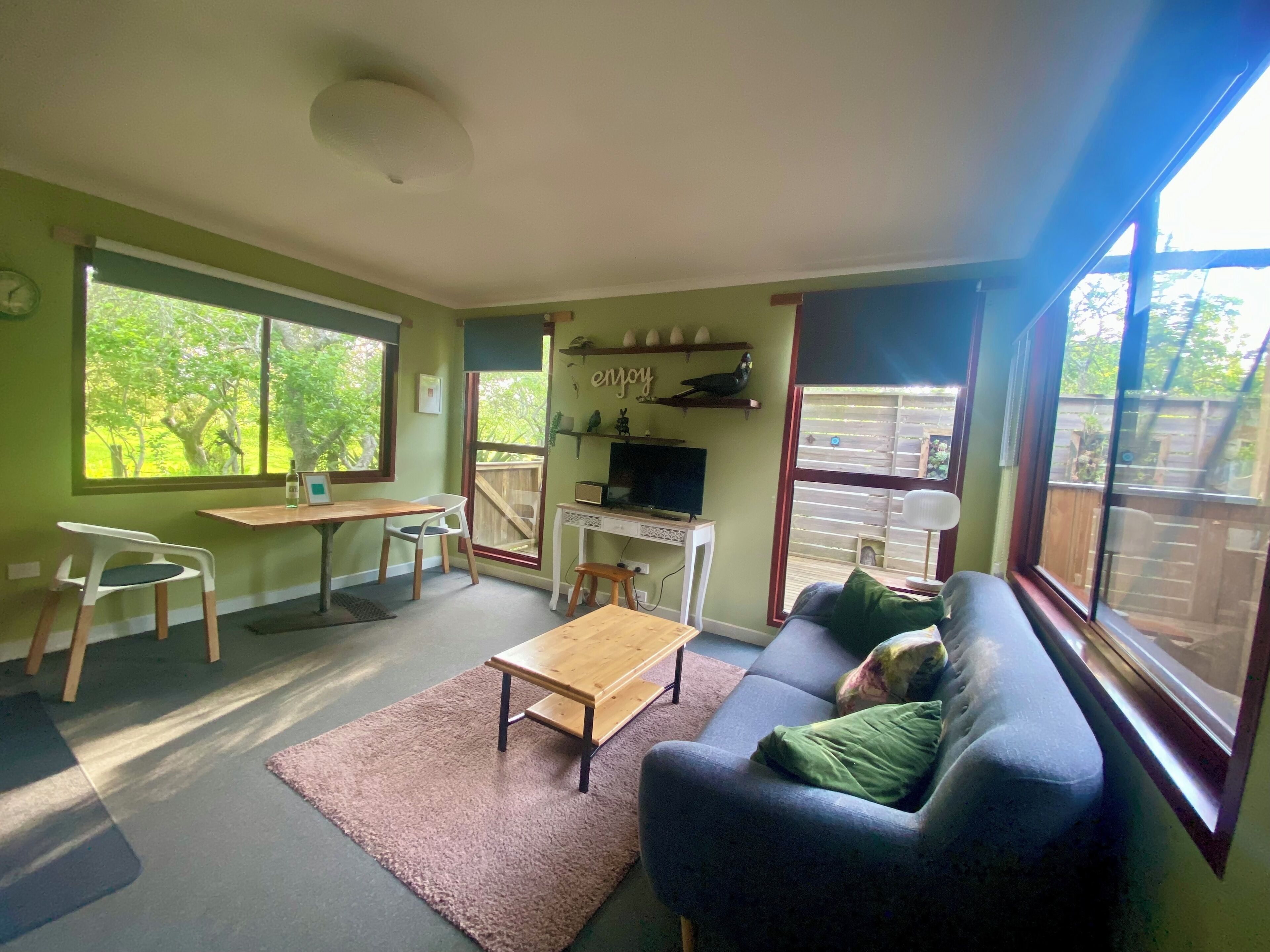 A living room with a sofa, a coffee table, a TV, and a desk with a chair. There are large windows and a view of greenery outside.