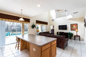 A spacious living area with a kitchen island, a brown leather sofa, a flat-screen TV, and a staircase leading to an upper level.