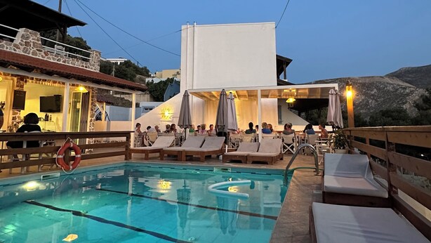 A swimming pool with lounge chairs and umbrellas, surrounded by buildings with outdoor seating and a mountainous backdrop. - Mary Popi (Kalymnos)