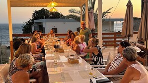 A covered outdoor dining area with a long table set for a group of people, overlooking the sea. - Mary Popi (Kalymnos)