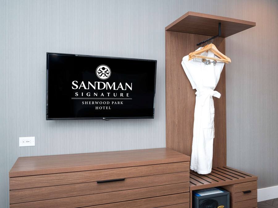 Habitación de hotel con un televisor de pared mostrando el logo del Hotel Sandman Signature Sherwood Park, un mueble de madera con cajones, una bata colgada y un microondas al fondo.