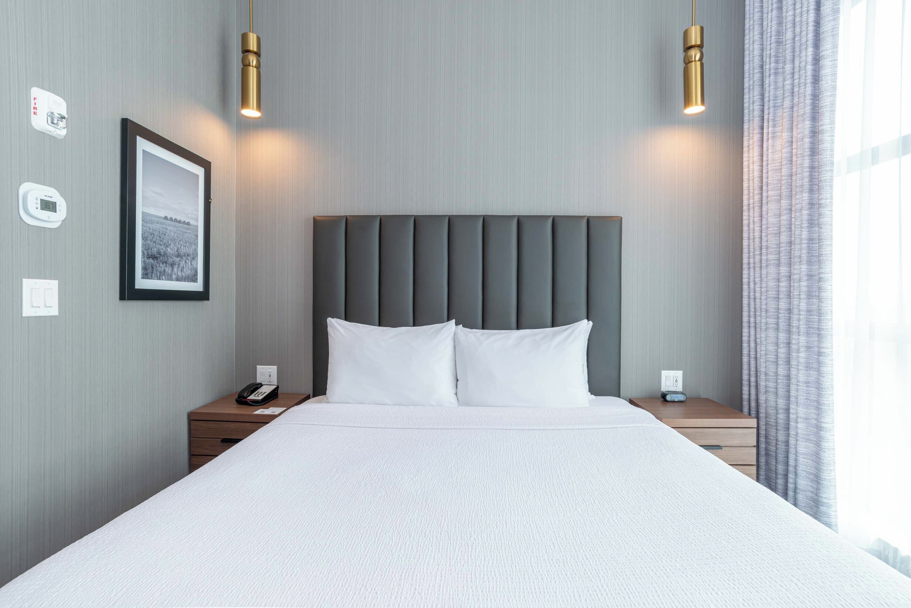Una cama bien tendida con un cabecero tapizado gris, dos mesitas de noche con lámparas, un cuadro enmarcado en la pared y una ventana con cortinas.