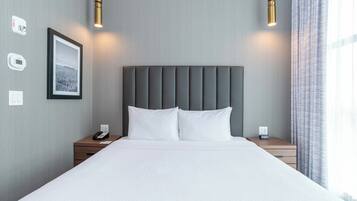 Una cama bien tendida con un cabecero tapizado gris, dos mesitas de noche con lámparas, un cuadro enmarcado en la pared y una ventana con cortinas.