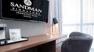 A hotel room with a bed, a desk with a coffee maker, a TV displaying the hotel name, and a window with curtains.
