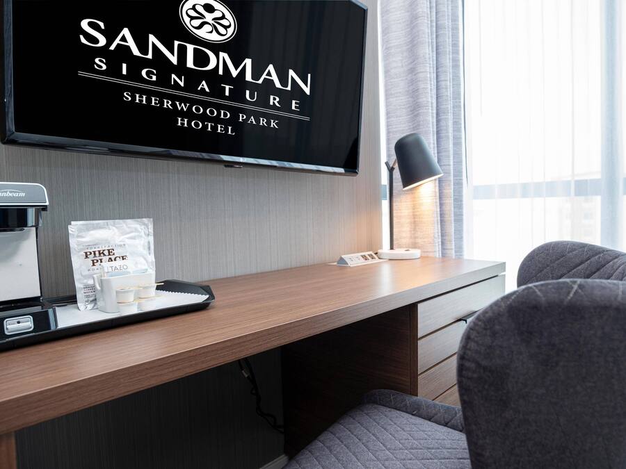 Una habitación de hotel con cama, escritorio con cafetera, televisor mostrando el nombre del hotel y ventana con cortinas.