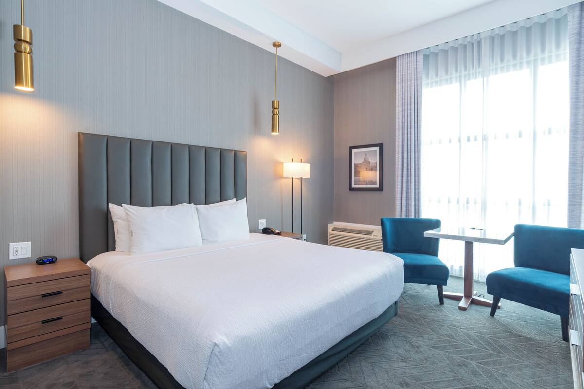 Een hotelkamer met een groot bed, nachtkastjes, een bureau, twee stoelen, een lamp en een raam met gordijnen.