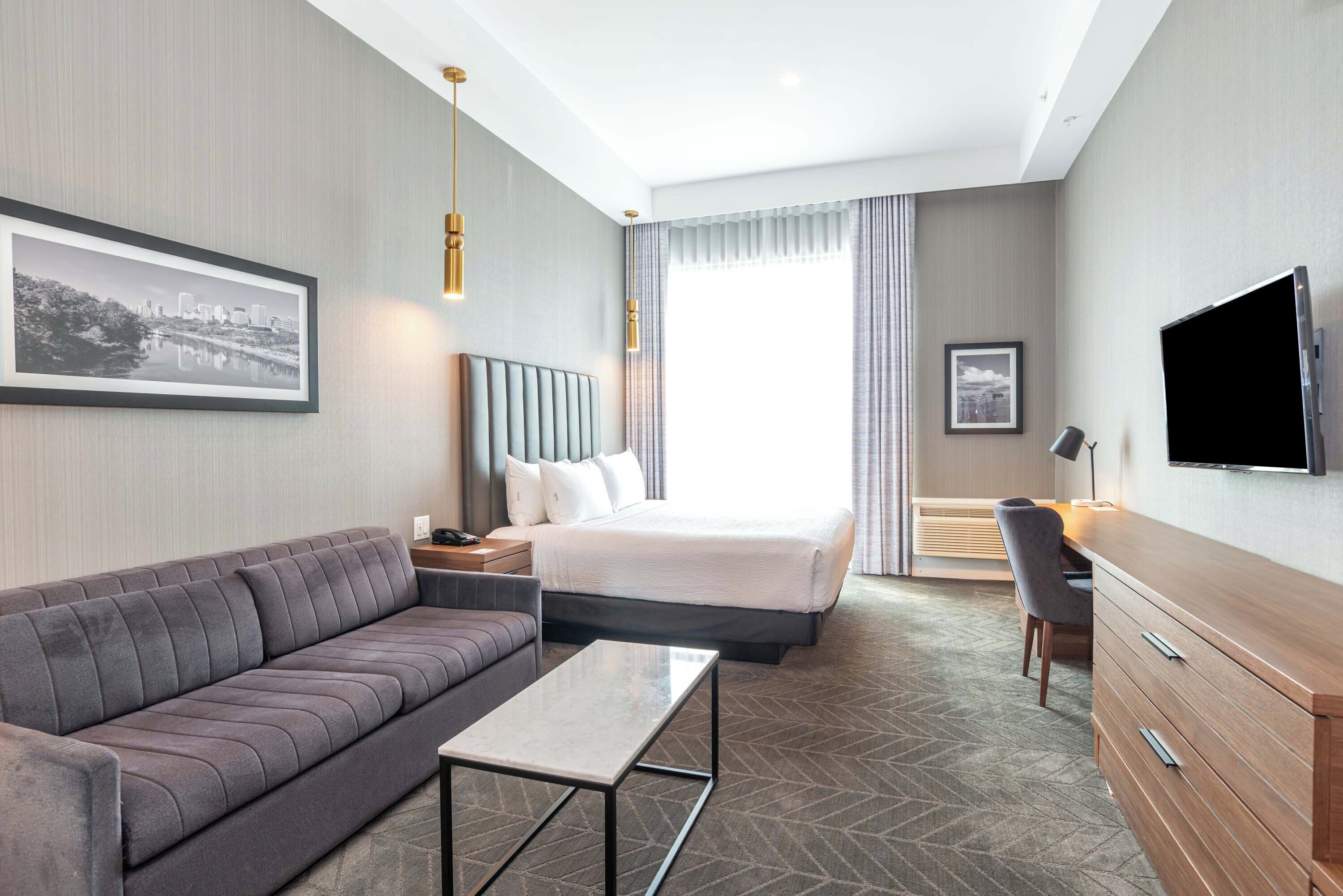 A hotel room with a bed, a sofa, a desk, a TV, and a framed picture on the wall.