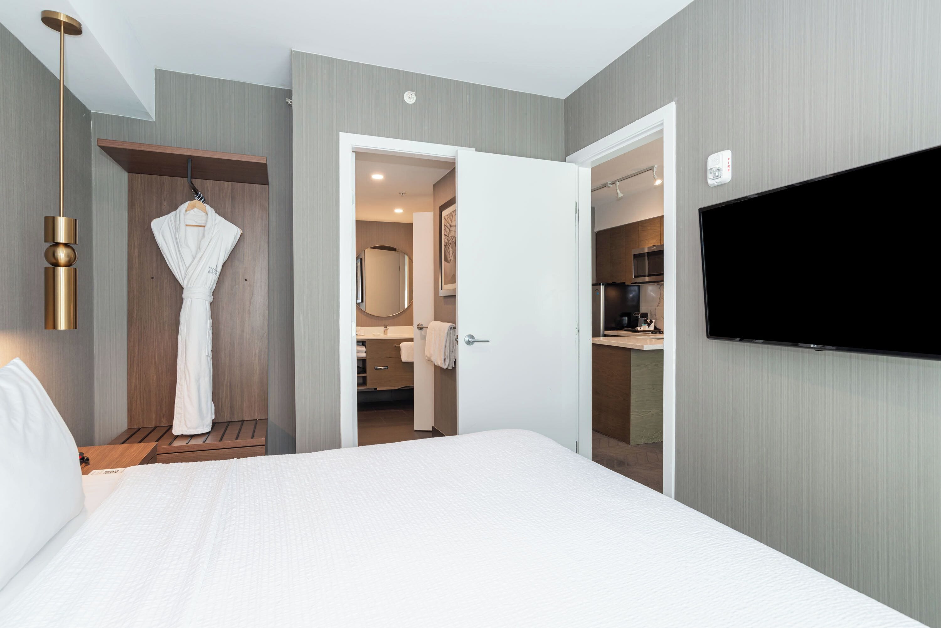A hotel room with a large bed, a robe hanging on a wooden shelf, a TV mounted on the wall, and a door leading to a bathroom.