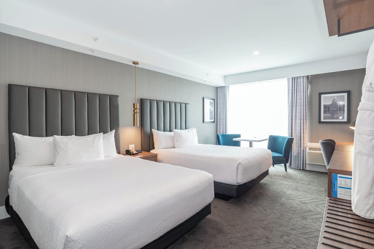 Een hotelkamer met twee bedden, een grijs hoofdeinde, een tafeltje, een stoel en een groot raam.