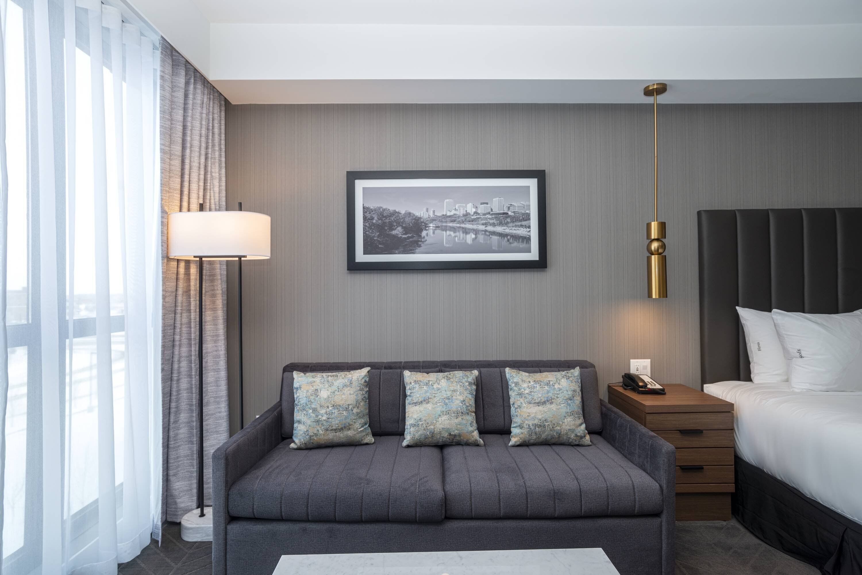 Una habitación de hotel moderna con un sofá gris, una cama, una mesita de noche, una lámpara y un cuadro enmarcado con una vista urbana.