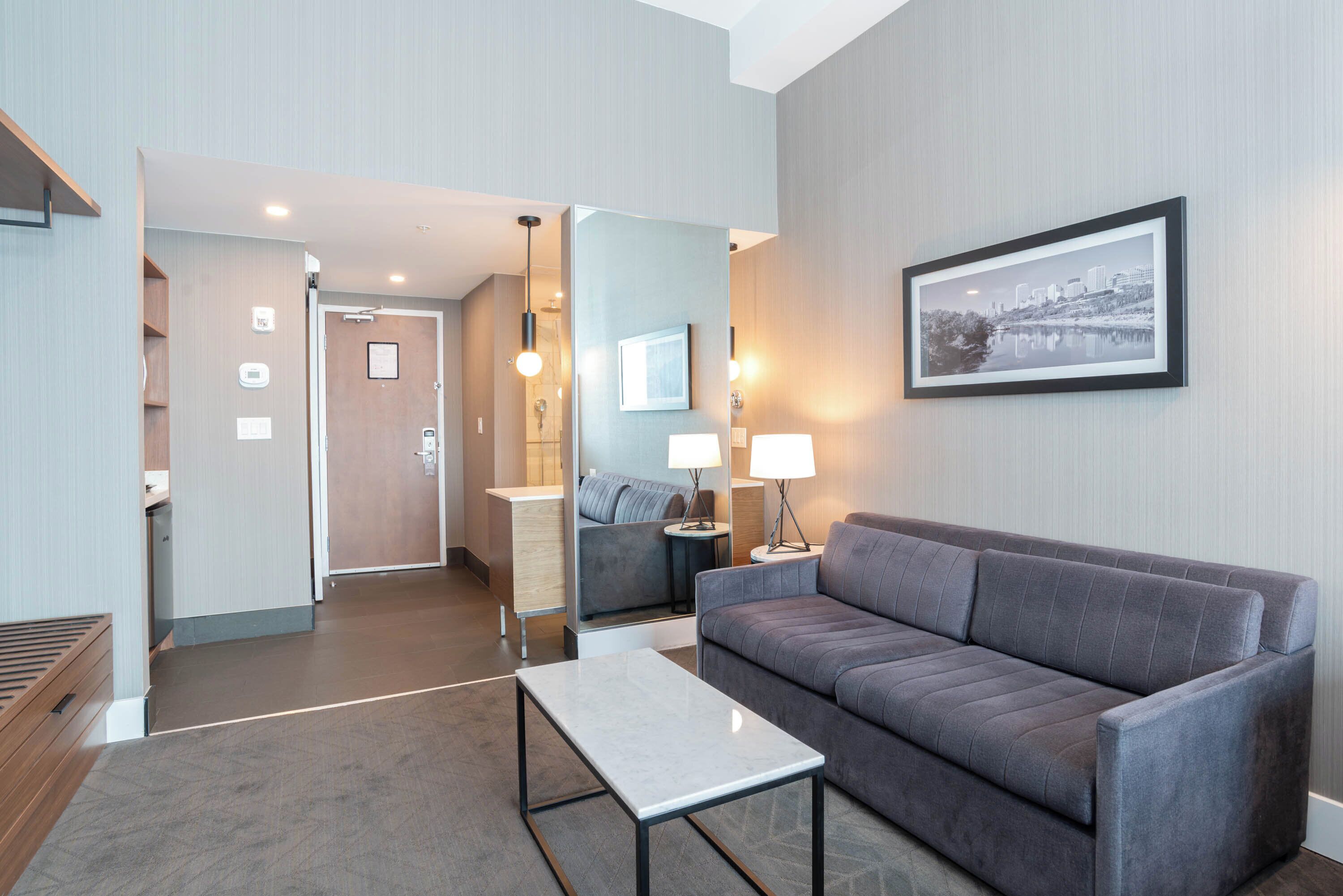 A modern hotel room with a grey sofa, a small table, a mirror, and a framed picture on the wall.