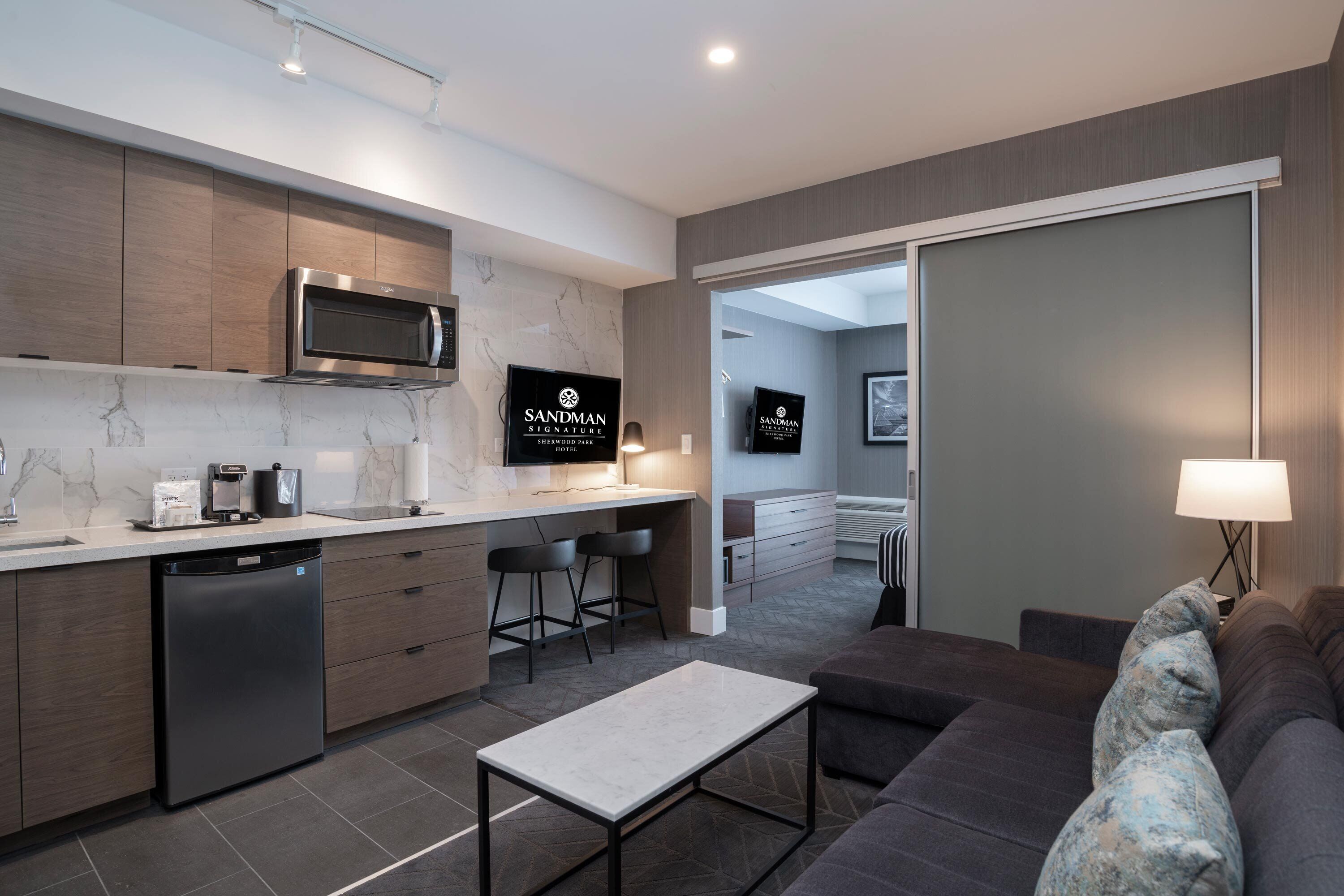 A modern kitchen with a microwave, dishwasher, and bar stools. There is a sofa and a lamp in the living area.
