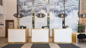 Een receptiegedeelte met drie receptiebalies, elk voorzien van een zwarte mat ervoor. Er zijn decoratieve wandpanelen en hanglampen.