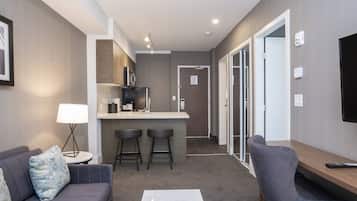 A modern hotel room with a sofa, a small table with a lamp, a bar area with stools, and a TV mounted on the wall.