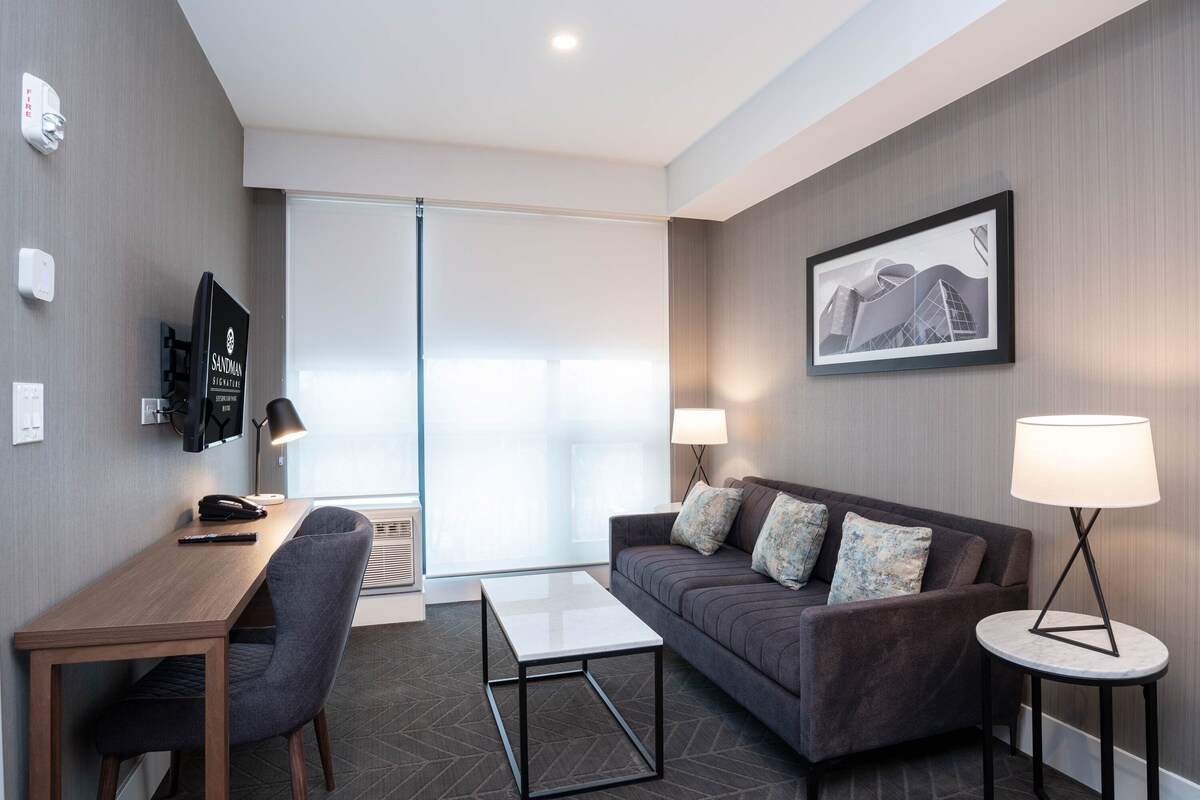 Une chambre d’hôtel moderne, équipée d’un canapé, d’une petite table, d’un bureau avec une chaise, d’une télévision et d’un tableau encadré au mur.