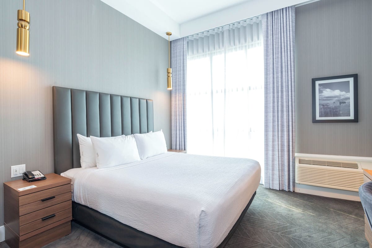 Een moderne hotelkamer met een groot bed, een nachtkastje met telefoon, een wandheater en een groot raam met gordijnen.