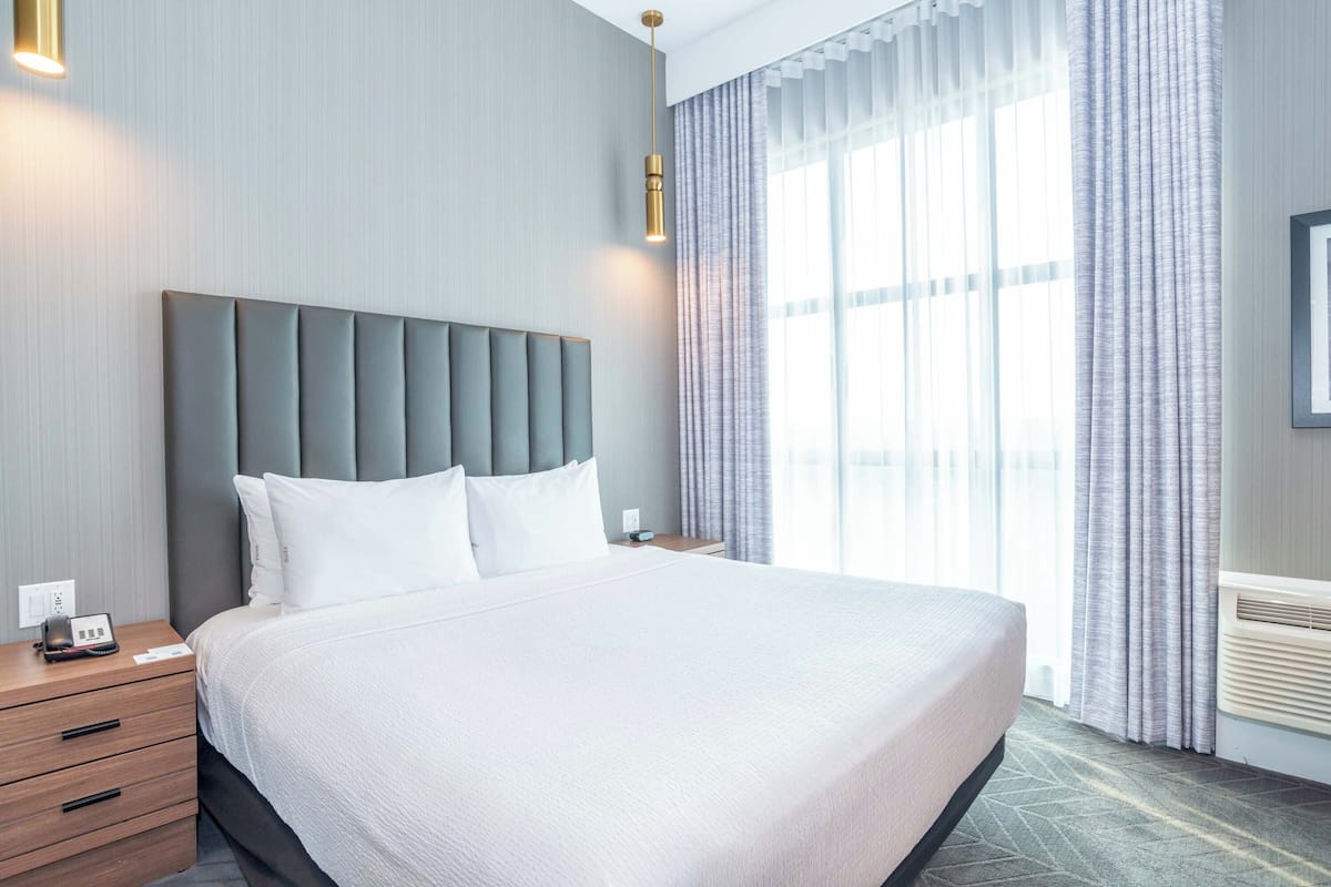 Een groot bed met een grijs bekleed hoofdeinde, witte beddengoed en een nachtkastje met een telefoon en een klein voorwerp erop.