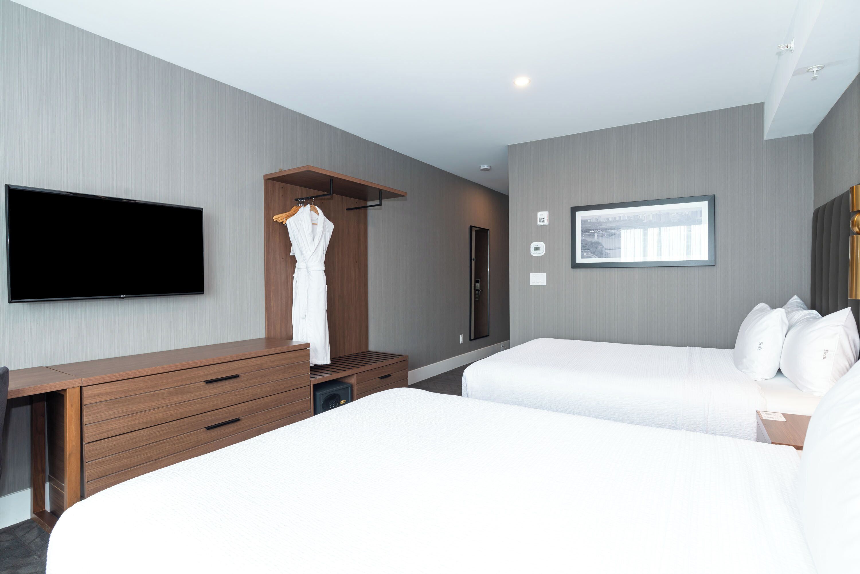A modern hotel room with a flat-screen TV, a wooden dresser, a robe hanging, two beds, and a framed picture on the wall.