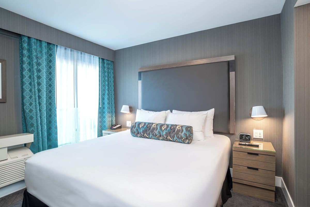 Ein Hotelzimmer mit einem großen Bett, Nachttischen, einem Beistelltischchen mit Lampe und einer Wanduhr.