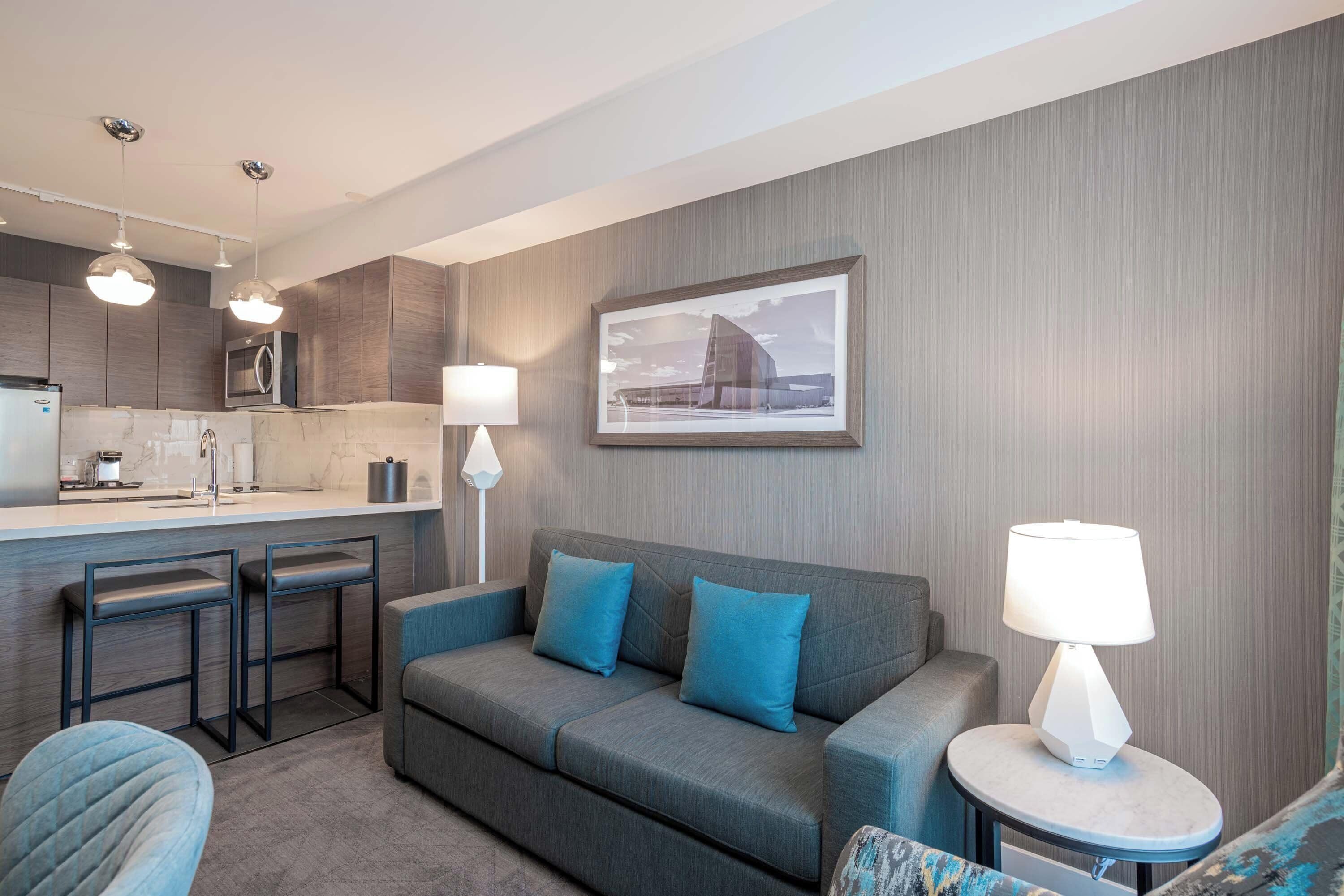A modern hotel room with a kitchenette, a grey sofa with blue cushions, a round table with a lamp, and a framed picture on the wall.