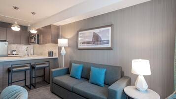 A modern hotel room with a kitchenette, a grey sofa with blue cushions, a round table with a lamp, and a framed picture on the wall.