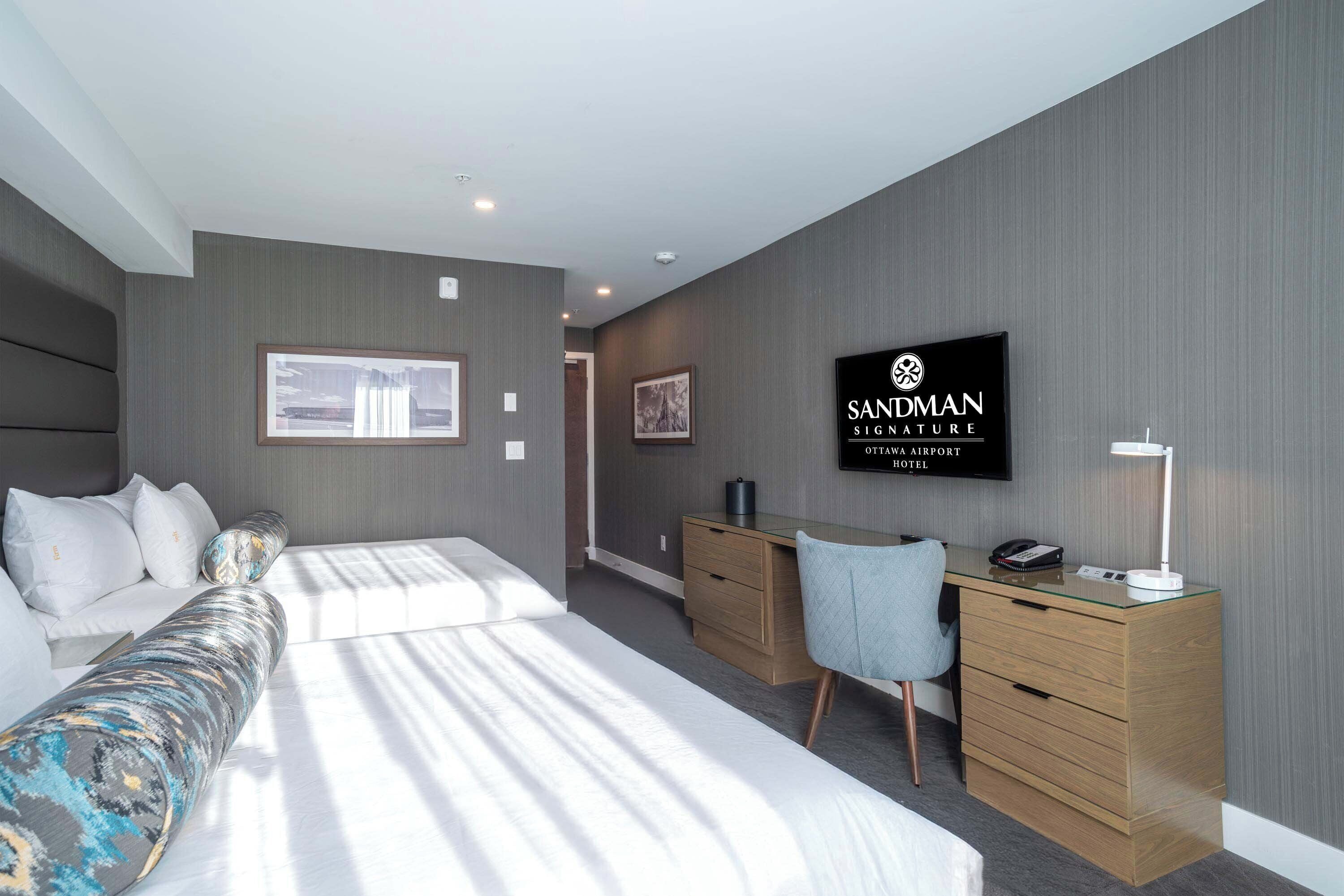 Habitación de hotel moderna con cama, escritorio, silla y televisión mostrando el nombre del hotel.