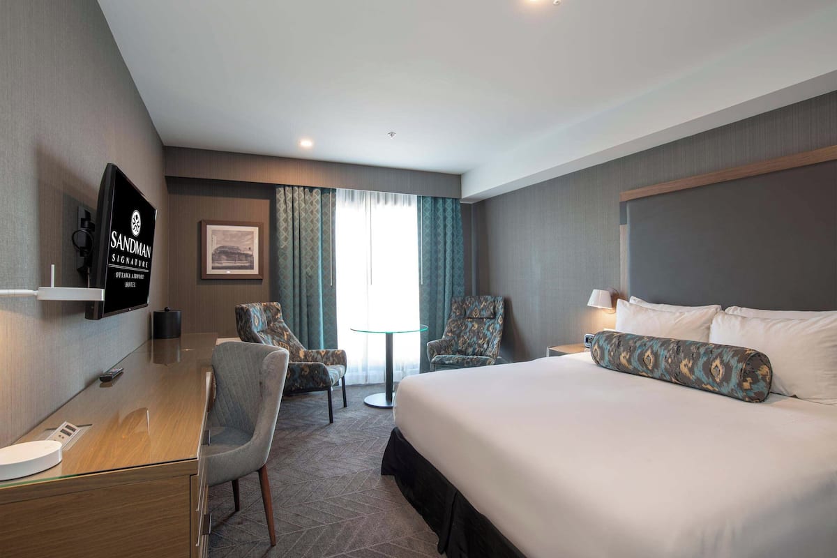 Ein Hotelzimmer mit einem Bett, zwei Sesseln, einem Schreibtisch mit Fernseher und einem fensterbepflanzten Fenster.
