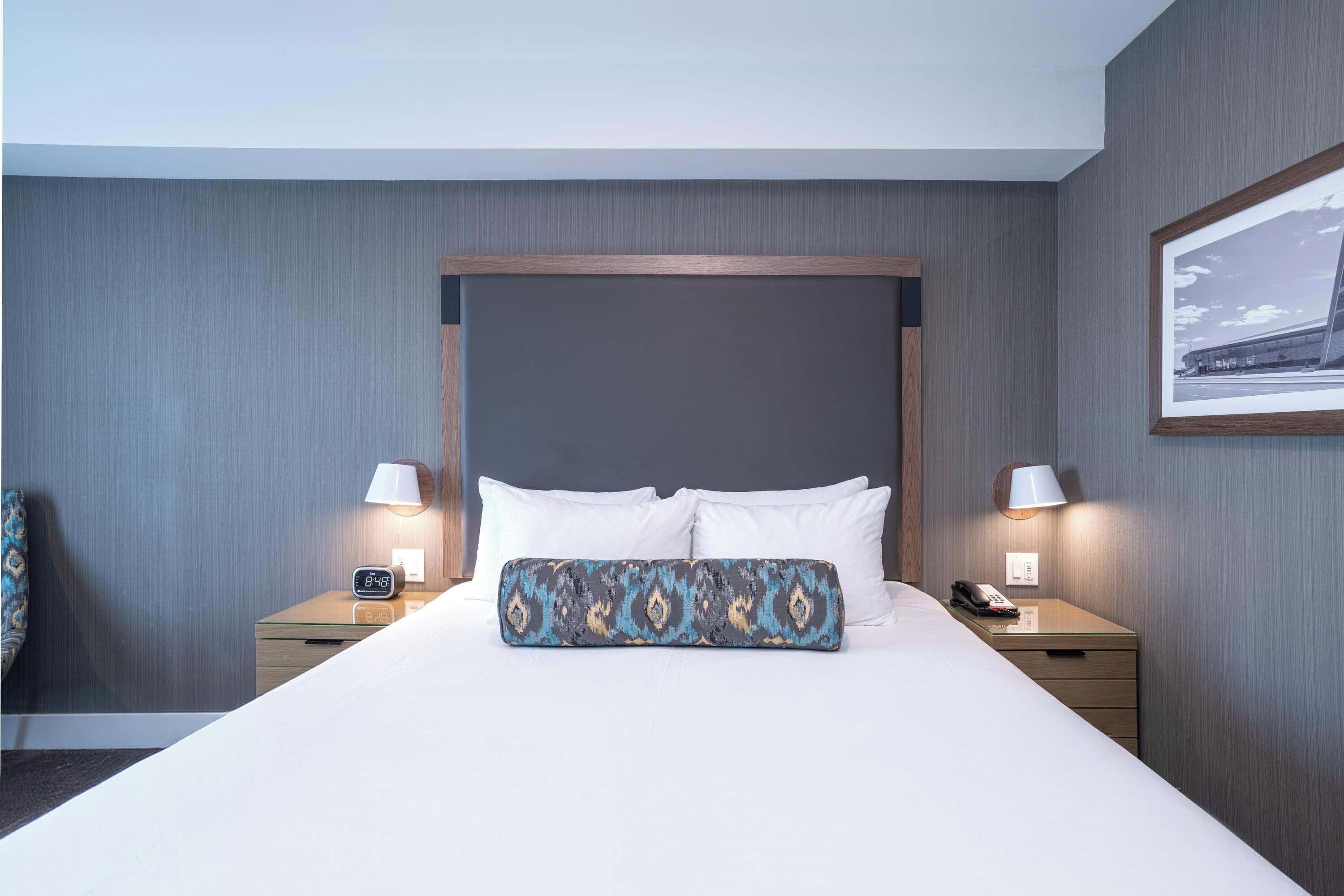 Ein Hotelzimmer mit einem großen Bett, zwei Nachttischen, einer Lampe, einer Uhr und einem gerahmten Bild an der Wand.