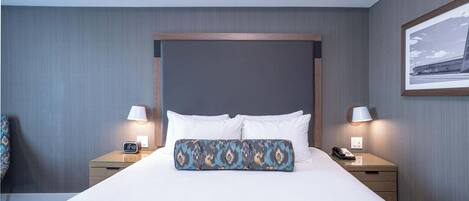 A hotel room with a large bed, two bedside tables, a nightstand, a lamp, a clock, and a framed picture on the wall.