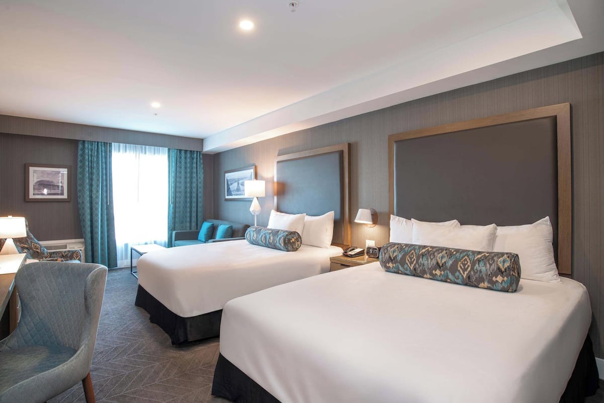 Ein Hotelzimmer mit zwei Betten, einem Schreibtisch, einem Stuhl und einem großen Fenster mit Vorhängen.