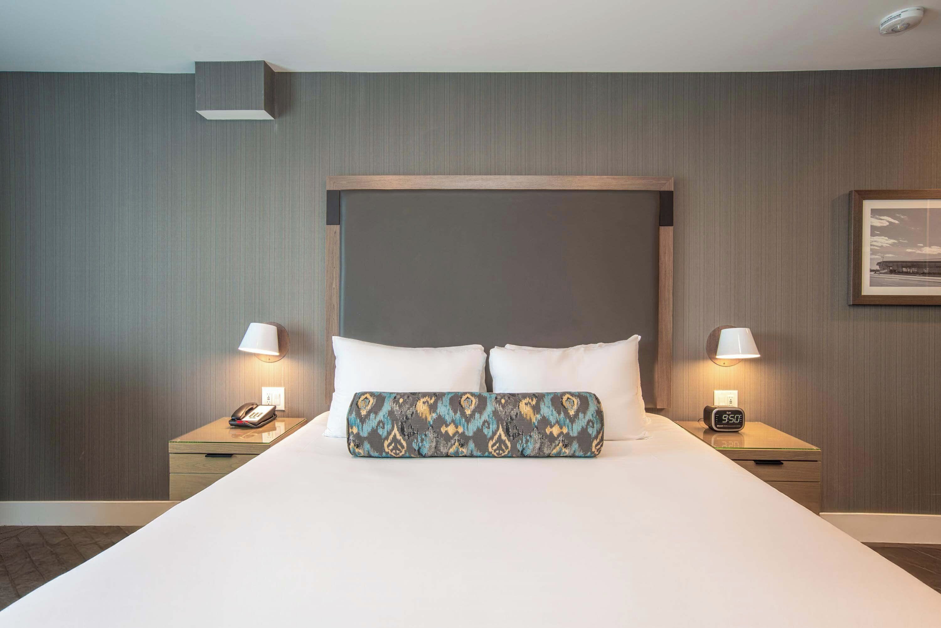 Una habitación de hotel con una cama grande, dos mesitas de noche, una mesita de noche con lámpara y un cuadro colgado en la pared.