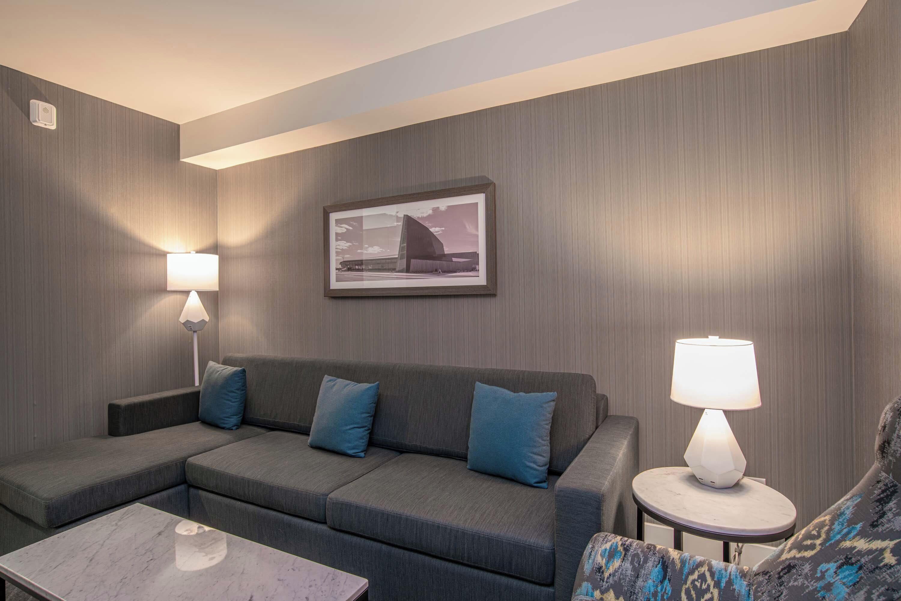 A modern hotel room with a grey sofa, two blue cushions, a small round table with a lamp, and a framed picture on the wall.