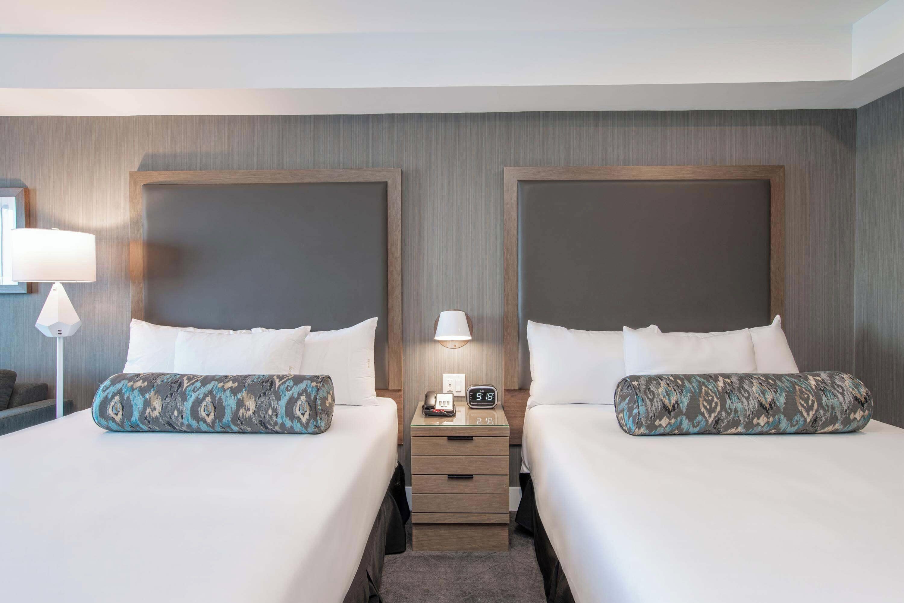 A hotel room with two beds, a bedside table with a lamp, and a nightstand with a phone and clock.