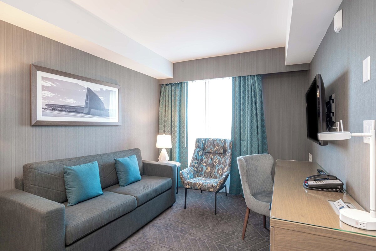 Une chambre d’hôtel équipée d’un canapé, d’un fauteuil, d’un bureau, d’une lampe, d’une télévision et d’un tableau encadré au mur.