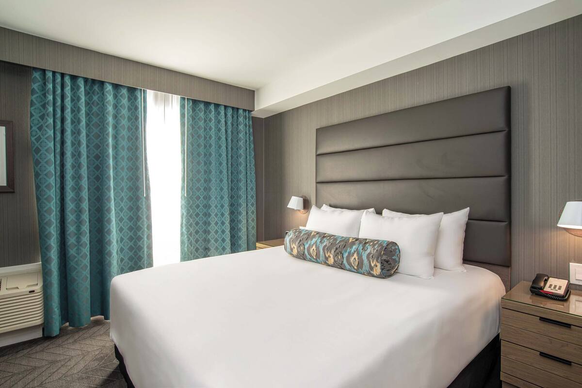 Ein Hotelzimmer mit einem großen Bett, einem Nachttisch mit Lampe, einem Telefon und einem an der Wand montierten Fernseher.