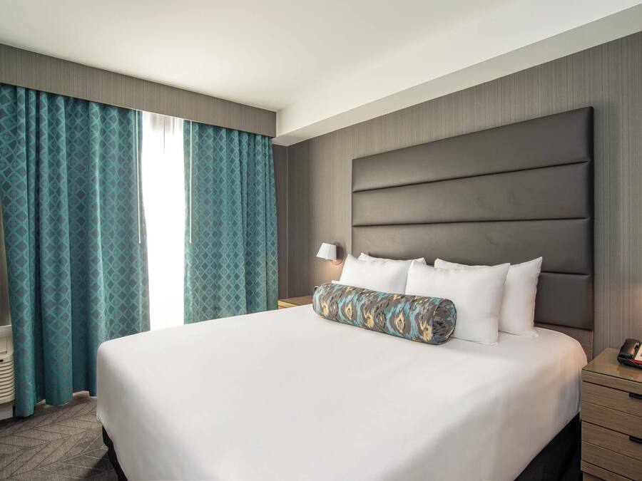 A hotel room with a large bed, a bedside table with a lamp, a phone, and a wall-mounted TV.