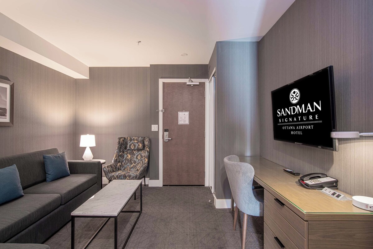 Une chambre d’hôtel avec un canapé, un fauteuil, un bureau, une télévision et une porte.