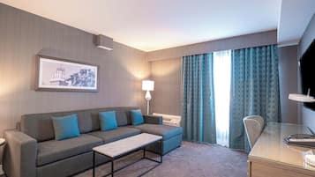 Habitación de hotel con sofá, mesa de centro, escritorio, lámpara y televisor.