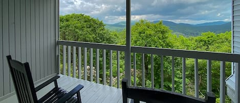 A wooden deck with a white railing, two wooden chairs, and a view of a mountainous landscape with trees.
