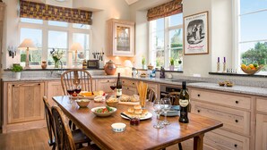 A well-lit kitchen with wooden cabinets, a dining table set with food and drinks, and large windows offering a view of the outdoors. - The School House, Coryton - a peaceful retreat  (sleeps 6+toddler) (Devon)