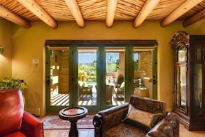 A room with a wooden ceiling, a sofa, a red chair, a table, and a view through glass doors to an outdoor area with a pool and greenery. - Los Valverde - Exclusive Luxury Home, Unsurpassed Views, Pool and Hot Tub! (Santa Fe)