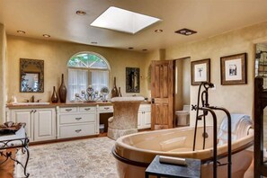 Ein geräumiges Badezimmer mit einer freistehenden Badewanne, einem Waschtisch mit Spiegel und einem großen Fenster.